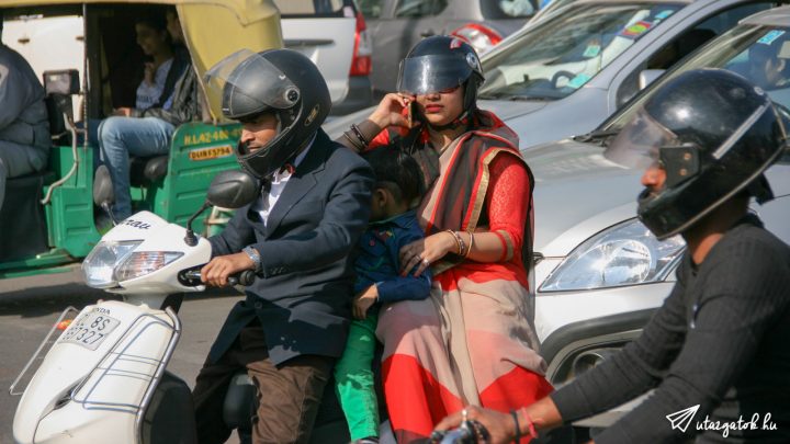 három indiai a motoron, elöl a férfi, hátul a nő, aki telefonál, köztük pedig valószínű a gyermekük