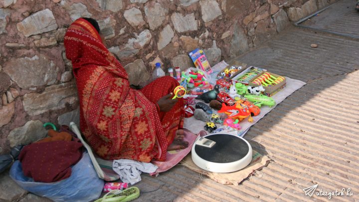 Indiai asszony árulja portékáit a földön, előtte egy fürdőszobai mérleg