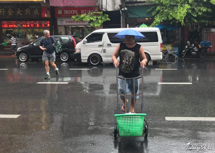 Thai férfi a sapkájára erősített esernyővel, így mindkét keze szabadon marad, amivel egy kocsit tol